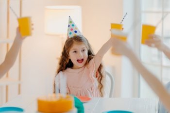 Happy ginger girl wears party hats, clink cups with drink, has fun with friends, celebrate birthday together, pose in decorated room at home. Children, celebration and holiday concept
