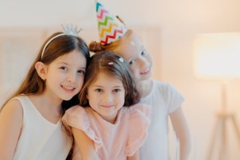 Portrait of happy three friends wear festive clothes, party hat, pose indoor against white background, play together during birthday celebration. Cute girls have good mood, come on special occasion