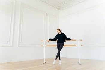 Classic ballet dancer has stretching workout, stands near ballet barres, wears black sweatshirt and leggings, does exercises in studio, focused down. Certified female instructor of choreography