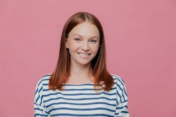 Headshot of pretty smiling European woman with charming smile, wears striped jumper, has brown hair, looks directly at camera, isolated over pink studio background. People, joy, happiness concept