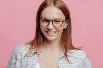 Photo of attractive cheerful young woman with glad satisfied expression, brown hair, smiles broadly, wears spectacles, delighted with new job, works in bank, meets with colleagues, poses indoor