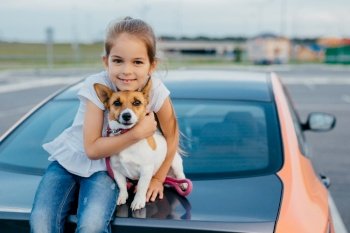 Little girl with appealing appearance, hugs her favourite pet, have journey together with parents by car, sit at trunk, pose for making photo. Children, animals, rest and transportation concept