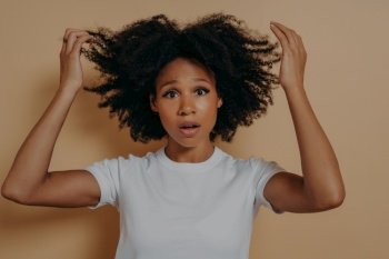 Worried young african american girl in white tshirt posing while standing isolated on beige studio background. Raising hands up in disbelief with shocked facial expression. Human emotions concept. Worried young african american girl in white tshirt posing on isolated beige studio background