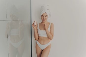 Image of young girl posing with cosmetic moisturizing face cream or serum in hand isolated over light wall in bathroom, wears towel wrapped on head after morning shower. Beauty and skincare concept. Young girl posing with cosmetic moisturizing face cream or serum in hand isolated over light wall in bathroom