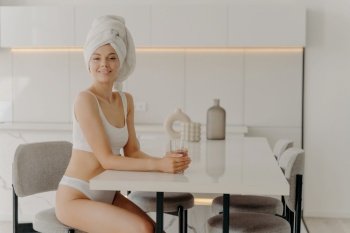 First thing in the morning. Relaxed slender woman sitting in stylish kitchen after taking shower with glass of pure mineral water, wears white underwear and towel over wet hair. Weightloss and dieting. Relaxed beautiful slender woman sitting in kitchen after taking shower with glass of water