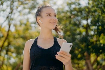 Thoughtful cheerful woman with pony tail has walk outdoor listens music while going in for sport holds modern smartphone leads healthy lifestyle focused into distance poses at park against trees