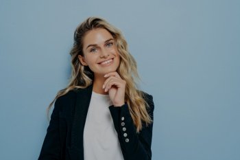 Young cute blonde woman touching with hand her chin, looking straight while smiling with big smile, making joyful and positive expression, posing isolated on blue background with copy space for text. Young blonde woman with hand on her chin smiling at camera
