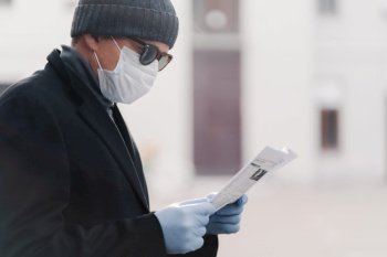 Cropped shot of man wears facial medical mask and protective rubber gloves, prevents coronavirus infection, reads newspaper, poses outdoor against blurred background. Quarantine, Covid-19 concept