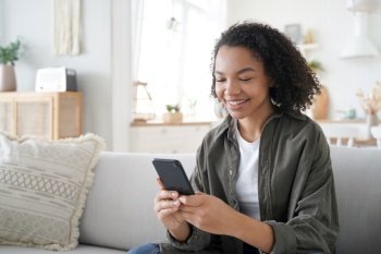 Curly african american girl has fun with phone at home. Happy hispanic woman is browsing social media and smiling. Chatting with friends remotely. Messaging with classmates. Communication concept.. Curly african american girl has fun with phone at home and smiling. Communication concept.
