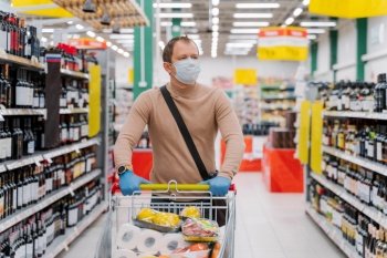 Horizontal shot of man walks in supermarket with shopping trolley with goods, wears medical mask and rubber gloves, food running out because of coronavirus. Virus outbreak. Panic buying concept.