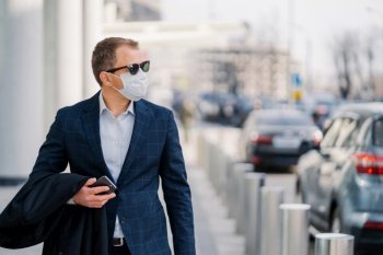 Thoughtful entrepreneur in formal wear, sunglasses, protects himself with sterile medical mask against coronavirus disease, walks outdoor, holds mobile phone, thinks about safety. Responsible behavior