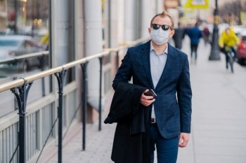 Outdoor shot of businessman in formal clothes, holds modern cell phone in hands, walks on street, protects himself with medical mask, prevents transmissible infectious disease or air pollution in city