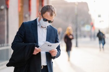 Coronavirus disease. Serious male banker reads newspaper attentively, finds out news about pandemic situation around world, wears protective mask, prevents spreading virus, stands at street.