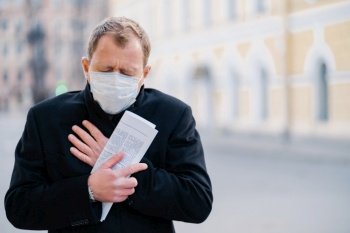 Infected man wears medical mask, has constant coughing, symptoms of Covid-19, holds rolled up newspaper, poses at city outdoor, needs isolation to stop spreading Coronavirus. Preventive measures