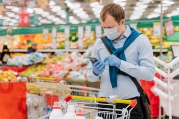 Young man poses in grocery store during coronavirus pandemic, wears protective medical mask and gloves, stands in supermarket near trolley, checks something in smartphone. Health, safety, quarantine