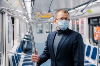 Male commuter poses in empty subway carriage, wears medical mask to prevent infected coronavirus at public metro station, looks somewhere, thinks about social distancing. Disease prevention.