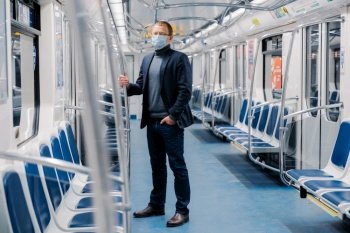 Full length shot of male entrepreneur in formal suit, medical mask, stands in urban train during disease outbreak, virus protection and quarantine protects himself from coronavirus at public transport