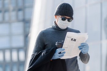 Flu disease, virus spreading protection. Young man wears medical mask against influenza viruses or coronavirus, poses in public space, reads press with concentrated look. Health care concept
