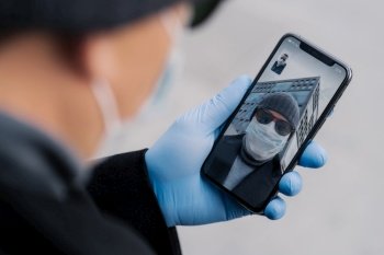 Shot of man has video conference via modern smartphone, being on distance from people during coronavirus spreading, wears protective mask and rubber gloves while walks outdoor in public place