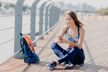 Outdoor shot of delighted pretty female sits crossed legs, uses modern cell phone for listening music and messaging with friends, takes break after jogging exercise, uses free internet connection