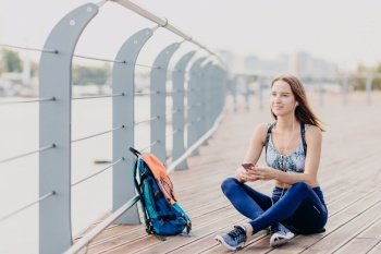 Beautiful relaxed sporty woman dressed in sportswear, sits crossed legs outdoor, enjoys pleasant music in earphones, focused into distance, uses modern technologies. People and rest concept.