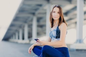 Photo of pleasant looking attractive sporty woman sits crossed legs, dressed in top and leggings, holds modern cell phone, enjoys lively music in earphones, poses outdoor. Entertainment concept