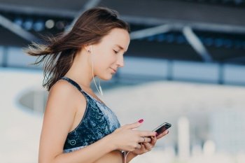 Sideways shot of sporty lovely female with dark hair, dressed in casual outfit, holds modern smart phone, checks notification, listens music with earphones, enjoys high speed internet connection