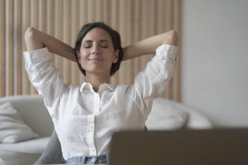 Attractive positive young female entrepreneur sitting at desk at home in relaxing position with hands behind her head and eyes closed, resting between training sessions while working remotely online. Positive female entrepreneur sitting at desk at home in relaxing position with hands behind her head