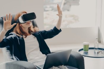 Businesswoman in virtual reality glasses using VR goggles, testing innovative method for business, sitting on armchair with opened laptop, interacting with digital interface. Cyberspace experience. Businesswoman in virtual reality glasses testing innovative method for business, working remotely