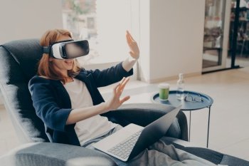 Confident young businesswoman wearing VR headset glasses and laptop sitting in cozy armchair in office lobby during her coffee break, raised hands up in air mimicking touch gestures in virtual reality. Young businesswoman wearing VR headset glasses and laptop sitting in cozy armchair in office lobby