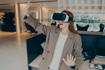 Surprised female office worker wearing VR headset surprised with how realistic virtual reality is, cannot contain emotions while testing 3d goggles at work, holding hands in front of her and gesturing. Woman wearing VR headset surprised with virtual reality while standing near her workplace in office