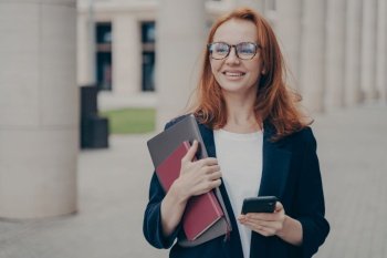 Happy red-haired business lady in official clothes standing outdoors, using mobile phone, being satisfied with work done after working day in office, holding notebook and smartphone in hands. Happy red-haired business lady in official clothes standing outdoors, using mobile phone