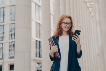 Happy european female holding mobile phone and smiling, reading good news from business partner while standing outside near office building with laptop, chatting with friend during coffee break. Happy european female holding mobile phone and smiling, reading good news from business partner
