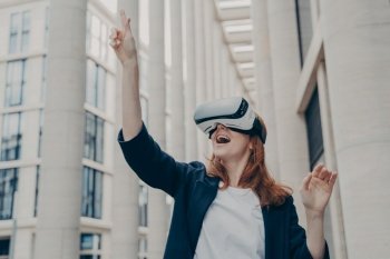 Augmented reality in business. Young excited happy female enterpruer wearing portable VR goggles trying to touch something in virtual reality with her finger, standing alone on city street. Excited female enterpruer wearing portable VR goggles trying to touch something in virtual reality
