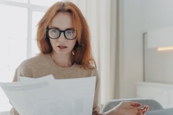 Serious redhead young woman focused in paper documents studies financial report wears spectacles casual jumper poses indoor against home interior. Busy ginger female financier does paperwork. Serious redhead young woman focused in paper documents studies financial report