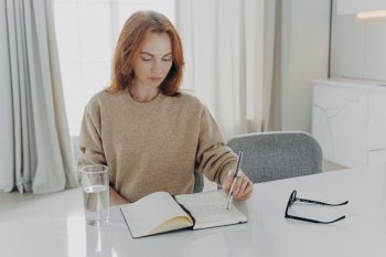 Indoor shot of serious redhead young woman makes notes in notepad makes list to do plans workday manager time sits at white desktop with glass of water spectacles against cozy domestic interior. serious redhead young woman makes notes in notepad sits at table