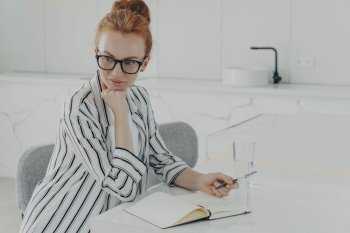 Young concentrated ginger woman in eyewear dressed casually thinking about new day while sitting at table in kitchen with transparent glass of water, looking aside while making notes in notebook. Young concentrated ginger woman thinking about new day while making notes in notebook at kitchen