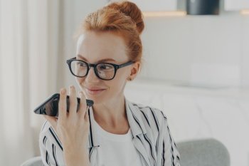 Horizontal shot of ginger woman uses voice assistant on smartphone makes voice call looks away has pleased expression wears transparent glasses casual striped shirt poses indoor. Technology.. Horizontal shot of ginger woman uses voice assistant on smartphone makes voice call