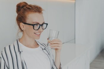 Indoor shot of smiling redhead young woman holds glass of water drinks aqua for body refreshment follows healthy lifestyle wears spectacles striped shirt focused into distance. Hydration concept. Smiling redhead young woman holds glass of water drinks aqua for body refreshment