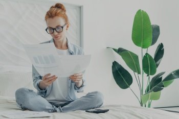 Casually dressed young woman in spectacles holding financial papers bills in hands while sitting on bed at home, having concentrated look while calculating family budget, house plant on background. Young focused redhead woman holding financial papers bills in hands while sitting on bed at home