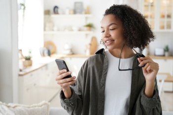 Curly african american girl has fun with phone at home. Young woman browsing social media and smiling. Chatting with friends remotely or watching video. Messaging with classmates and sharing content.. Curly african american girl has fun with phone. Messaging with classmates and sharing content.