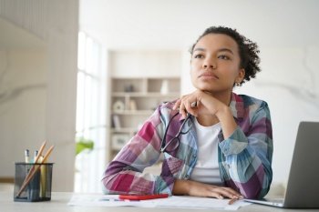 Teenage afro girl is bored at online lesson. Kid is distracted while studying remote. Teenage girl in front of laptop is looking far away. Remote study at high school or college at home on quarantine.. Teenage afro girl is bored at online lesson. Remote study at high school or college at home.