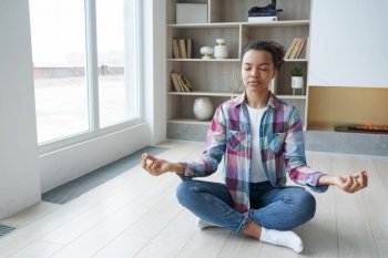 Hispanic young woman is sitting on floor in lotus pose and meditating. Teenage afro girl is practicing yoga at home and having fitness training. Concentration, zen and balance. Stress relief.. Hispanic young woman is sitting on floor in lotus pose and meditating. Home fitness training.