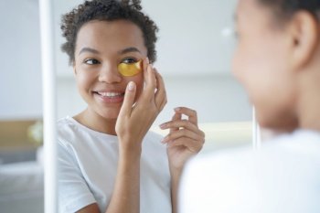 Mirror reflection of young hispanic lady. Happy afro girl is applying eye patches to her face. Skin moisturizer with collagen serum. Anti age patches. Morning skincare. Beauty routine of teenage girl.. Mirror reflection of young hispanic lady with eye patches. Skin moisturizer with collagen serum.