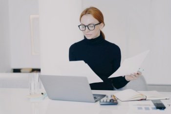 Concentrated woman secretary in office. Confident female entrepreneur or adviser at paperwork. White caucasian girl business lady is working at laptop and holding documents.. Concentrated woman secretary is working at laptop and holding document in office.