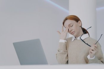 Employee has headache, touching her eyes. Forehead pain. Tired young woman at the desk in office. Girl is holding her head with hands. Concept of overworking and pressure at workplace.. Employee has headache, touching her eyes and forehead. Tired young woman at the desk in office.