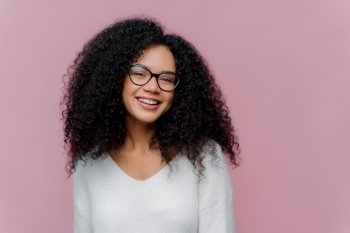 Headshot of pleased lovely woman with curly hairstyle, smiles gently at camera, wears optical glasses and white casual sweater, poses against purple background. Positive emotions and feelings concept