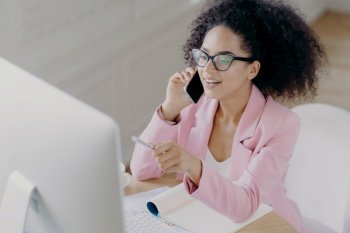 Glad young African American woman discusses business news, points with pen at screen on computer, talks via modern cell phone, busy with paper work, wears transparent glasses and formal wear
