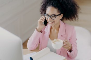 Top view of glad dark skinned woman with curly dark hair, has phone conversation, holds mug of drink, smiles pleasantly while looks at screen of computer, wears elegant attire, busy working at project