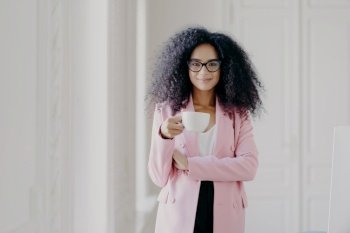 Shot of attractive woman with Afro hairstyle, drinks aromatic beverage, wears glasses, formal wear, poses in office over white background, copy space for your promotional content. People and business